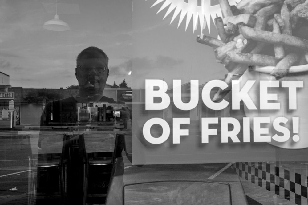 A man is reflected in a fast casual restaurant window.  A poster advertising a "bucket of fries" can be seen in the frame.