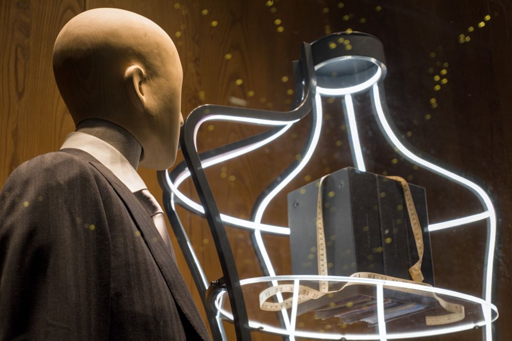 A mannequin in a store window appears to be staring longingly at a box with a measuring tape draped over it.  The box is sitting on a table which appears to be surrounded by neon lights.