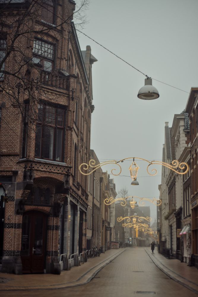 A photograph of a wet cobblestone street lined with historical commerce buildings. Overhead decorative lighting festively illuminates the misty scene. A pedestrian can be seen walking. Groningen, the Netherlands 