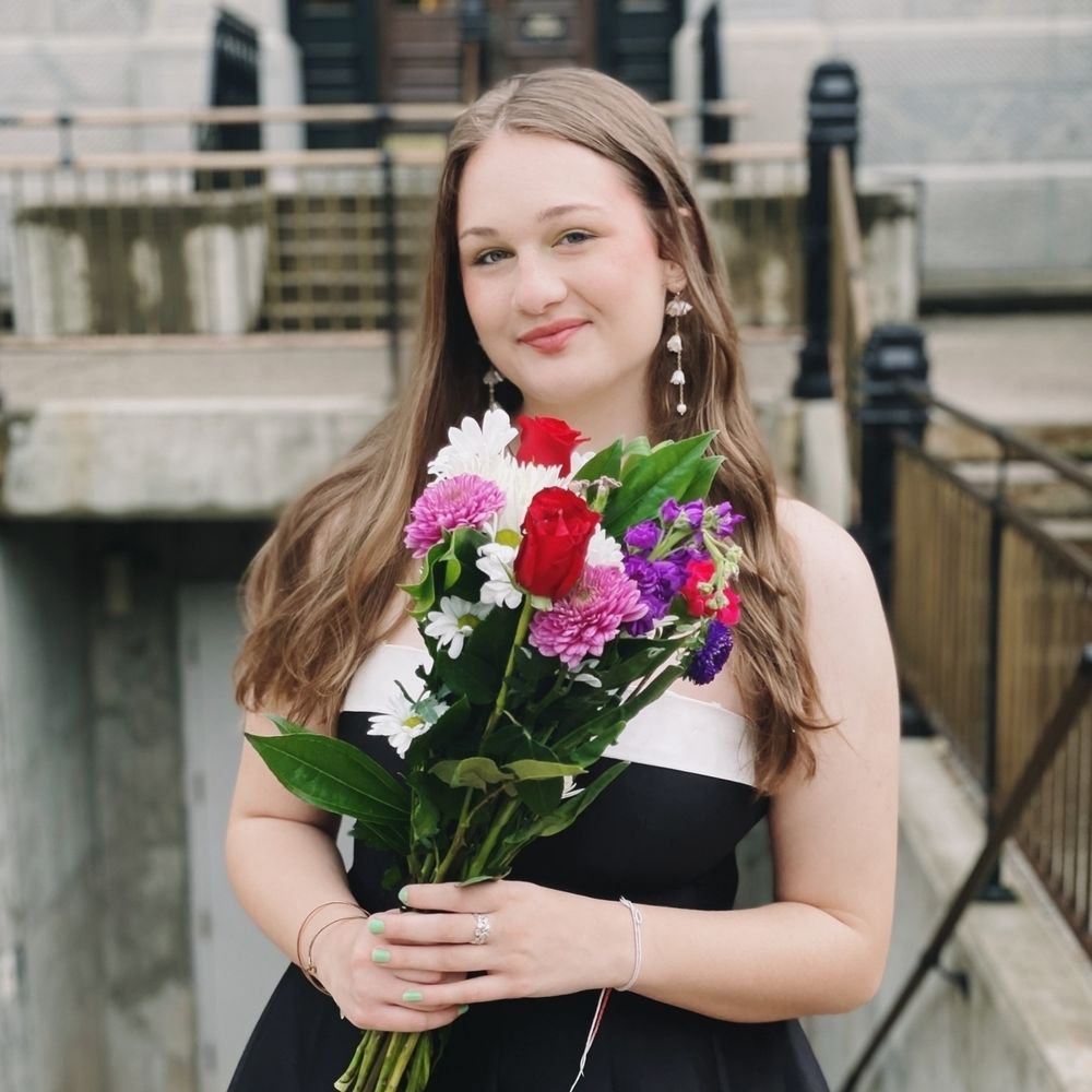 A person holding a bouquet of colorful flowers stands outdoors in a formal dress.