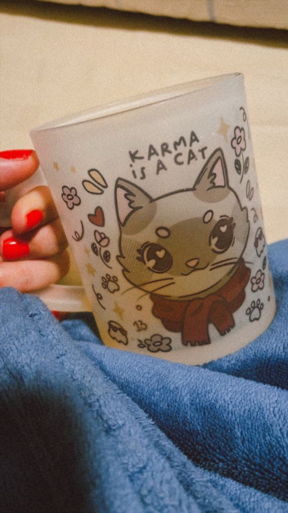 imagem de uma mão segurando uma caneca com cha. na caneca a imagem de um gato usando um cachecol vermelho. em cima do gato a frase “karma is a cat”. 