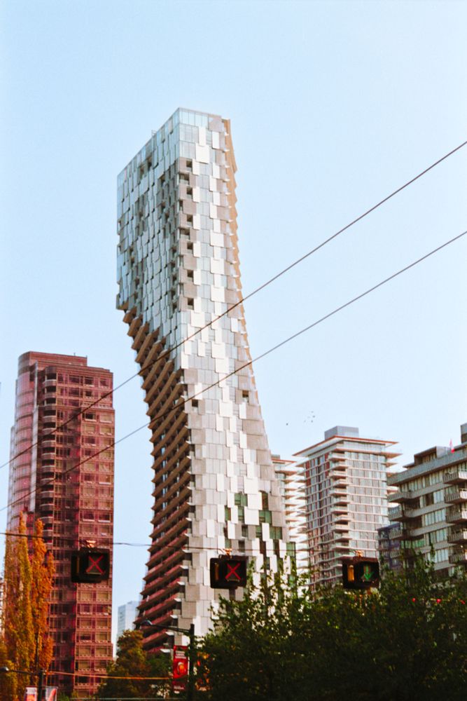 Those same three signal lights hang across the lower center of the frame, also two red X's on the left and middle and a green arrow on the right, but this time there are different buildings behind each one: a reddish/brownish postmodern highrise on the left, some more generic rectangular ones on the right, and in the middle is a stunning brand new tall glassy pillar that looks like someone took a bite out of the left side, all curved inward and back out in a semicircle down to the ground. The windows look like overlapping sheets of glass with no panes and the building slopes gently outward on the right side. Some trees fill the lower right foreground