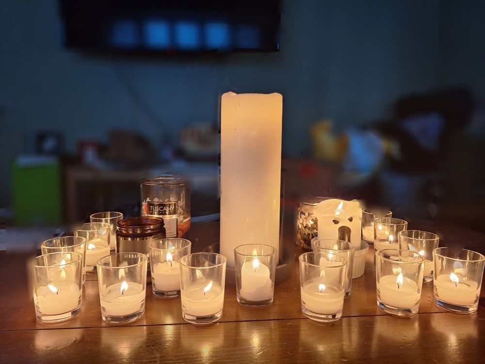 Many candles on a wooden table. Most are small white votive in glass jars, but there are also jar candles of various sizes and one oversized white pillar candle in the middle.