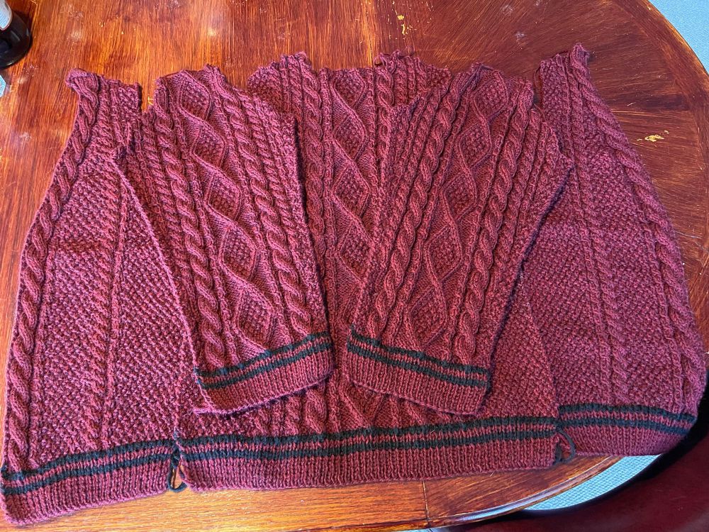 Five finished pieces of a hand knit cabled cardigan sweater ready to be seamed and finished. This picture highlights the sleeve design, which matches the back of the cardigan. The main color is garnet and the accent stripe color is black.