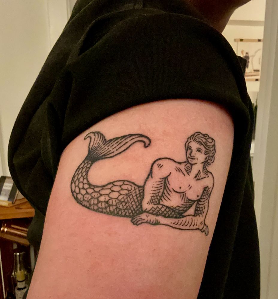medieval wood cut style tattoo depicting a pinup posed merperson with his tits out.