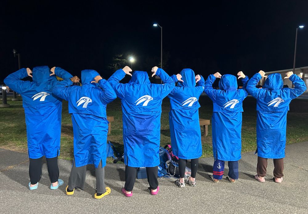 The back 6 girls standing in a row. All wearing royal blue long swim parkas, showing the Falcon logo on the back