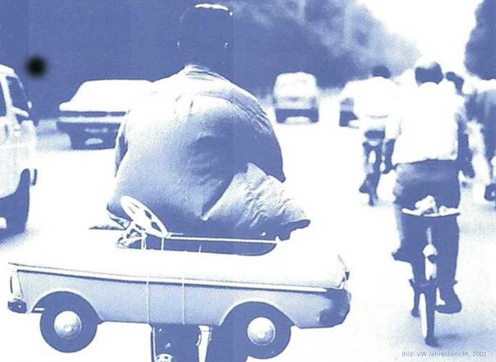 Ein Mann fährt Fahrrad und transportiert auf dem Gepäckträger ein Kinderspielzeugauto. Im Hintergrund sind weitere Radfahrende und Autos auf einer Straße zu sehen. Das Bild ist in Blautönen gehalten und spielt mit dem Kontrast zwischen Fahrrad und Auto.

A man rides a bicycle carrying a toy car on the rear rack. In the background, other cyclists and cars are visible on a road. The image is rendered in blue tones and contrasts the bicycle with the idea of motorized transport.