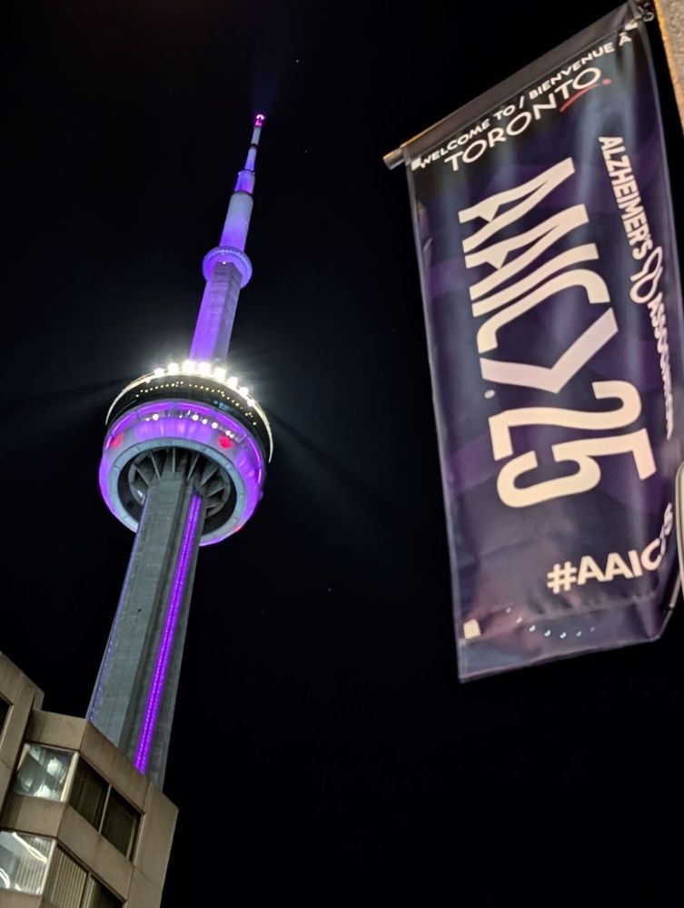 The CN tower shines purple in the night sky. The AAIC25 flag flies on a lightpole in the foreground.
