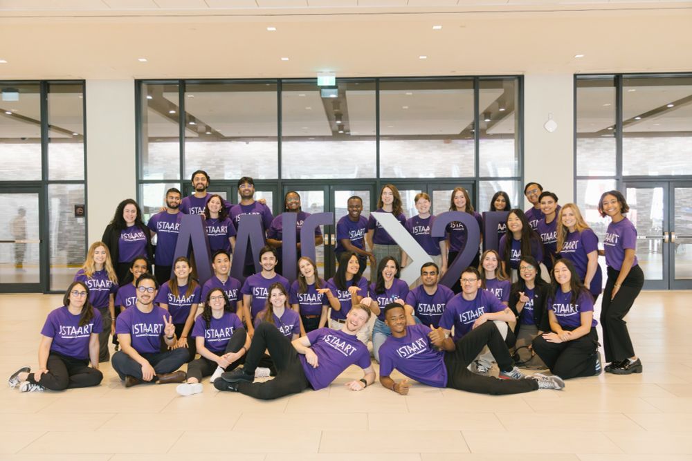 A large group of ISTAART members in matching purple t-shirts stand and sit around large AAIC25 letters, posing for a group picture.