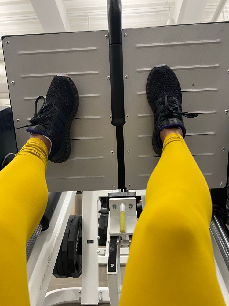 selfie shot of a woman’s legs on a leg press machine. woman is wearing mustard yellow leggings and black adidas 