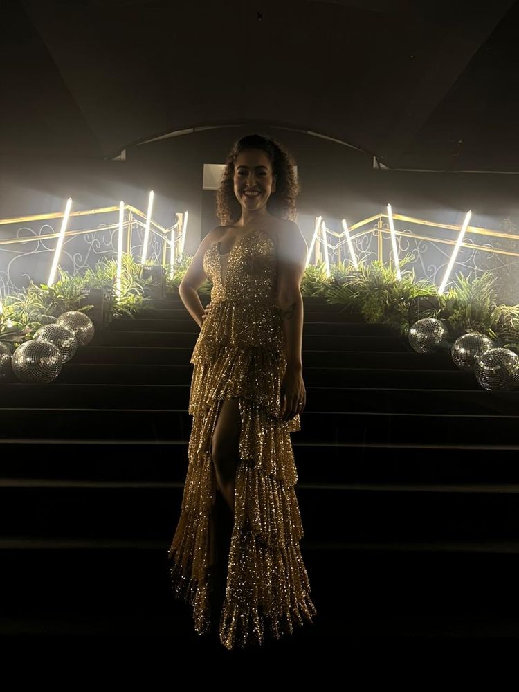 Foto na escada da primeira foto. A foto mostra o look completo iluminado pelas luzes ao fundo. O vestido cintila.