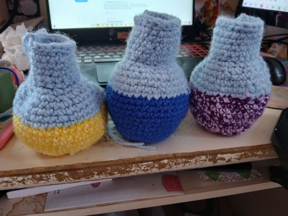 Three crocheted potion bottles; one in yellow, one in blue, and one in purple. They feature loose ends and no corks yet.
