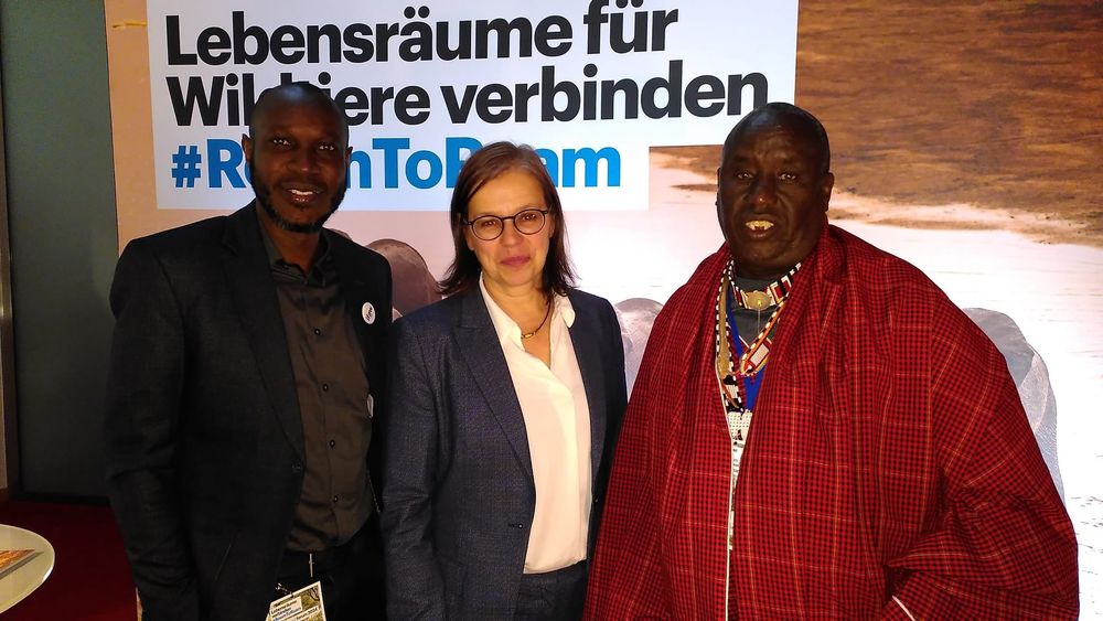 John Kogada und der Massai-Leader Daniel Leturesh stehen neben der Moderatorin Tanja Busse vor dem Schriftzug: Lebensräume für Wildtiere verbinden. 
