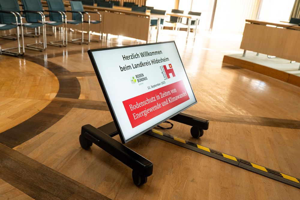 Sitzungsaal mit Holzboden darauf Monitor mit Anzeige: "Herzlich willkommen beim Landkreis Hildesheim. Bodenschutz in Zeiten von Energiewende und Klimawandel." 