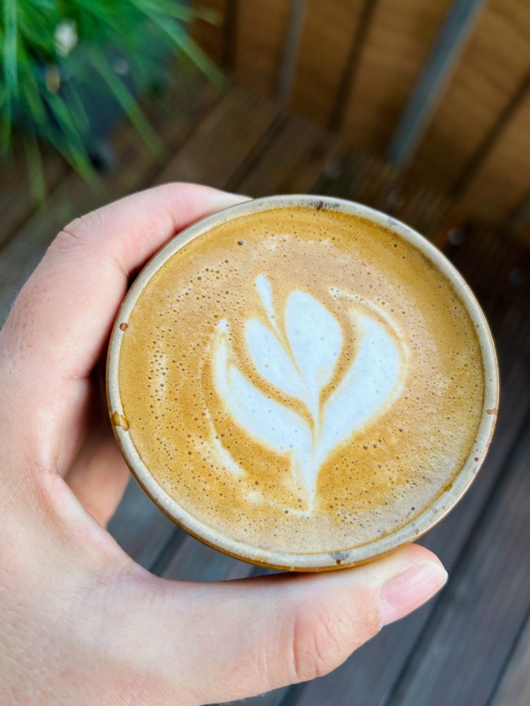 Tasse Kaffee mit Blumenmuster im Milchschaum. Balkonpflanzen im Hintergrund.