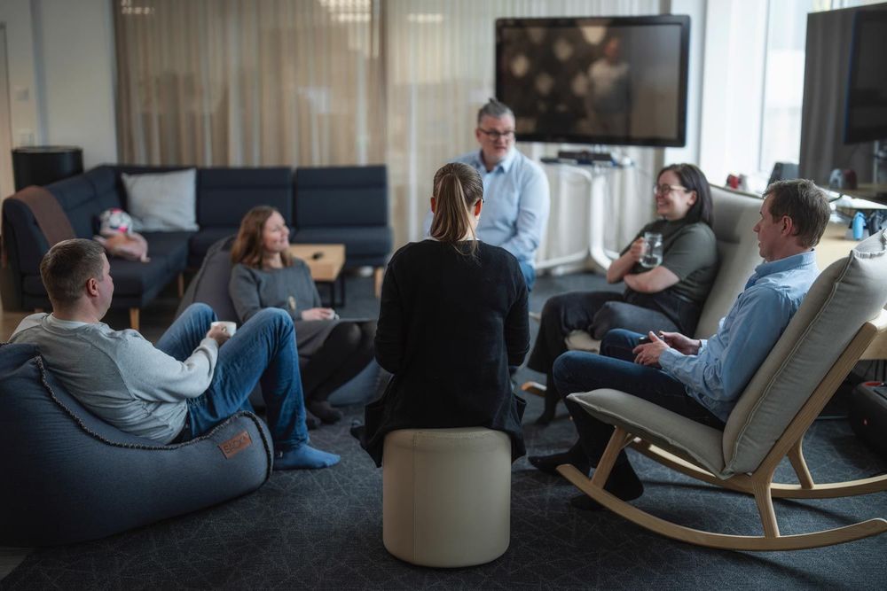 A group of six people are having a coffee break in the lounge area, sitting in a circle. They look relaxed, and the atmosphere looks cozy. 