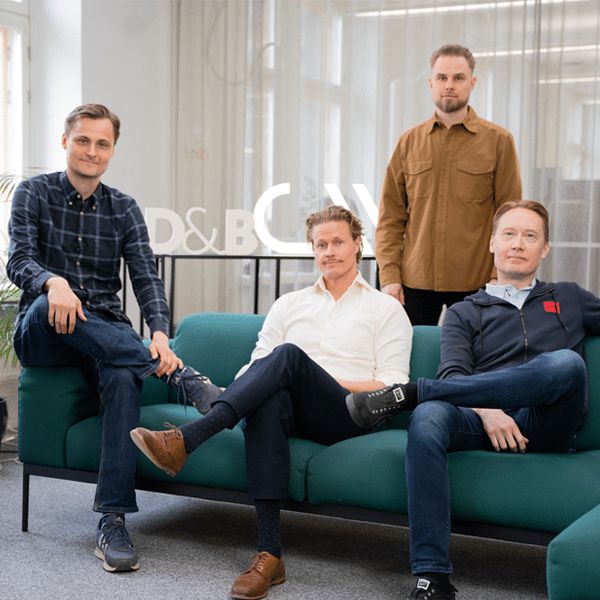 Four men pose for a group photo at Codemate's office. Three are seated on a green sofa, while one stands behind them. The setting is modern and light-filled, with a large window to the left and a translucent curtain in the background. All are dressed in smart-casual clothing and appear relaxed and professional, marking a business collaboration moment.