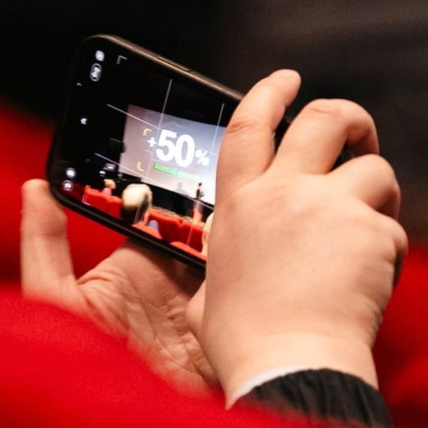 A person sitting in a red theatre seat captures a photo of a presentation on stage with their phone. The screen displays '-50%'