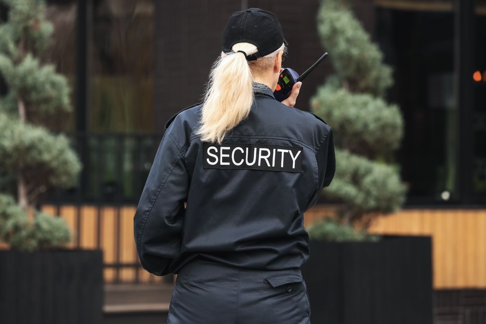 A female security officer with blonde hair tied in a ponytail, wearing a black uniform with 'SECURITY' written on the back. She is holding a walkie-talkie and appears to be monitoring the area outside a building with greenery in the background.