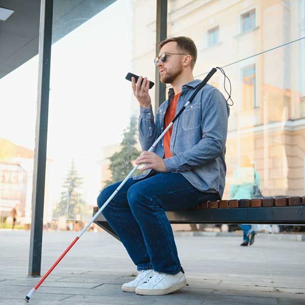 A blind man sitting at a bus stop bench, using a smartphone with voice input. He is holding a white cane and wearing sunglasses and casual clothes.