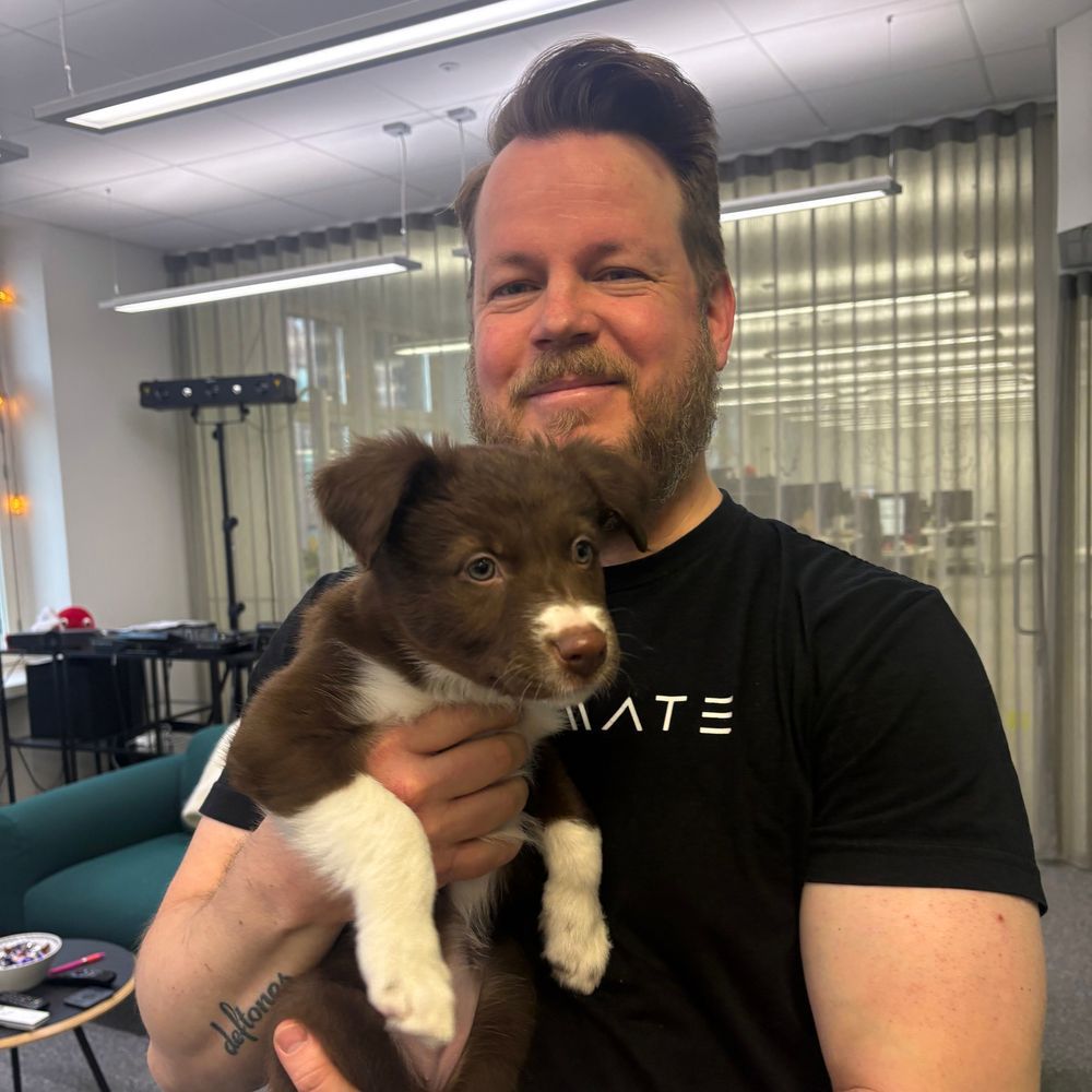 A man smiles while holding his brown and white border collie puppy in an office setting. He is wearing a black Codemate t-shirt and has a tattoo on his arm. In the background, there's a sofa, decorative string lights, and an open office space.