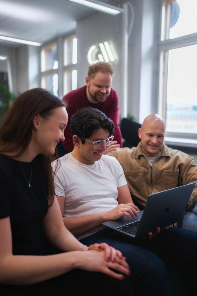 Four colleagues sitting closely together on a couch in a bright office, smiling and gathered around a laptop. One person is typing while the others look on, engaged and relaxed. A Codemate logo glows in the background.