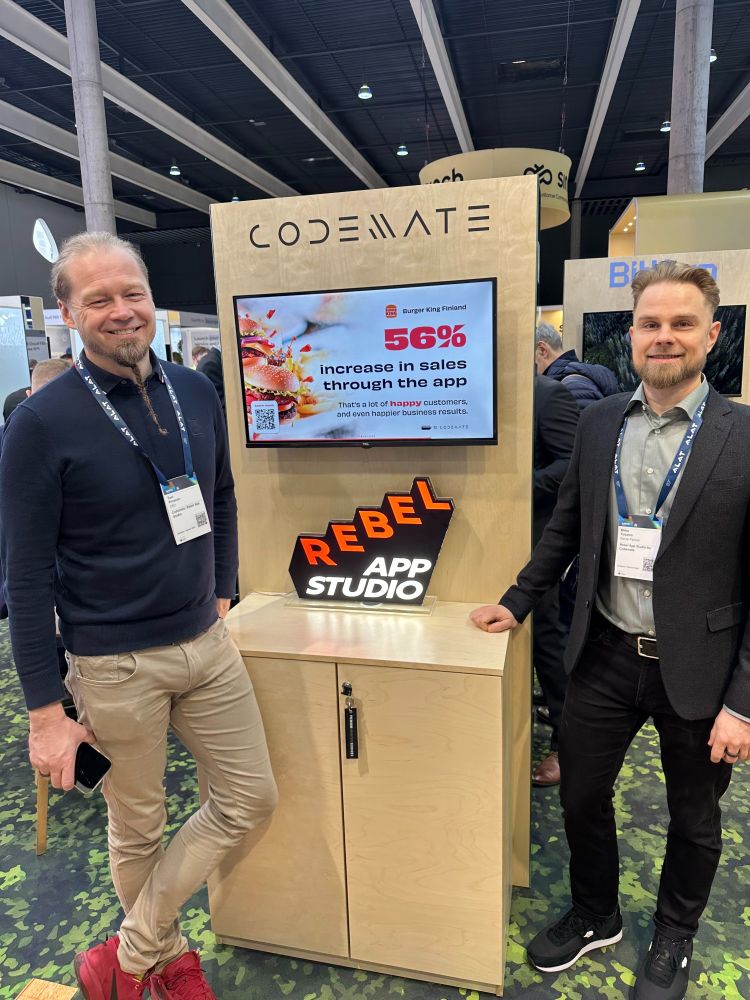 Miika and Toni standing at Codemate and Rebel App Studio’s booth at MWC25. A screen behind them displays a Burger King Finland case study showing a 56% increase in sales through the app. The booth features a wooden design with Codemate’s logo on top and a Rebel App Studio sign on the counter.