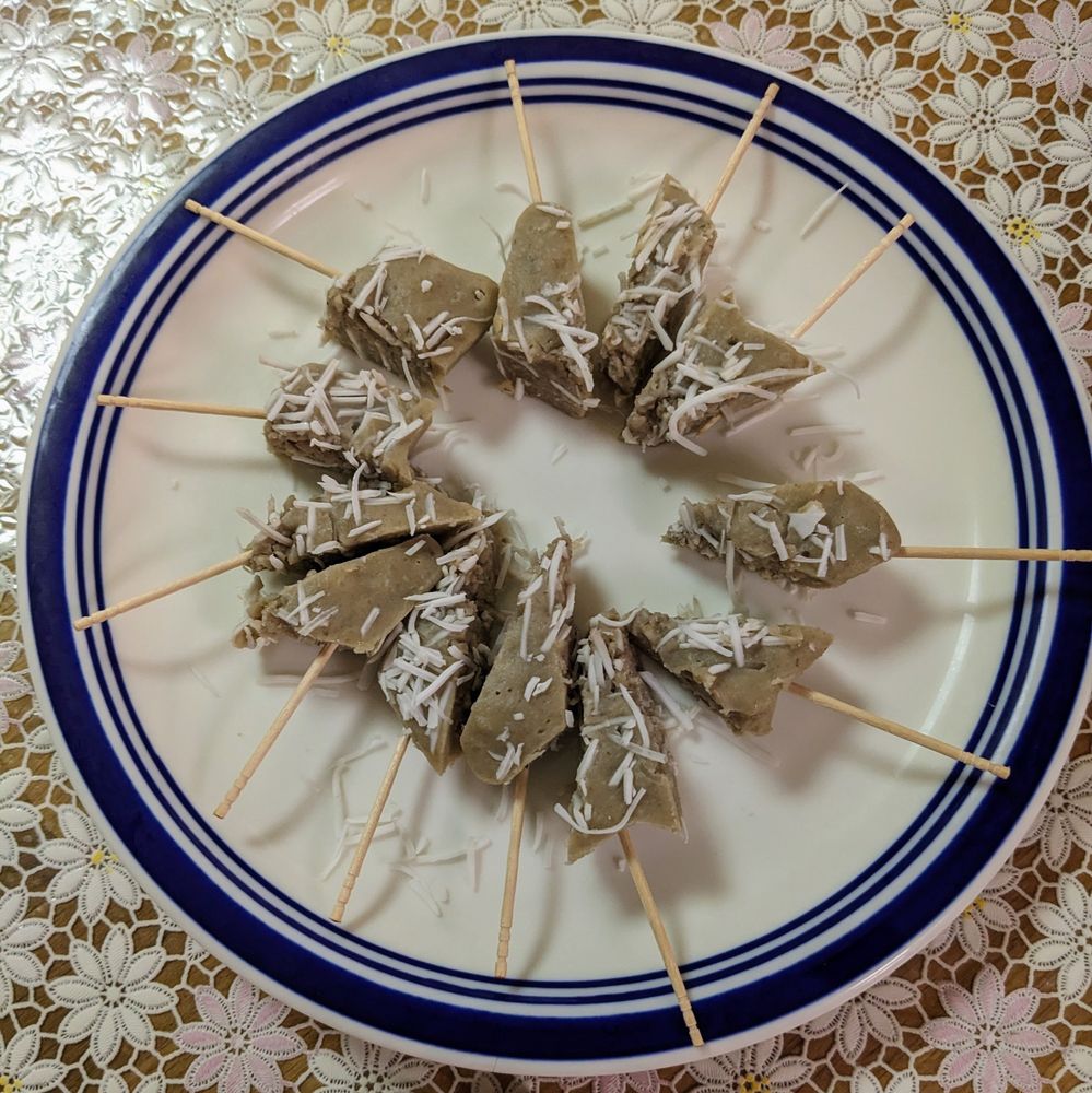 串に刺した三角形の茶色い蒸し菓子が円形に並べられ、上に白い細切りココナッツが散らされている白い皿の写真。