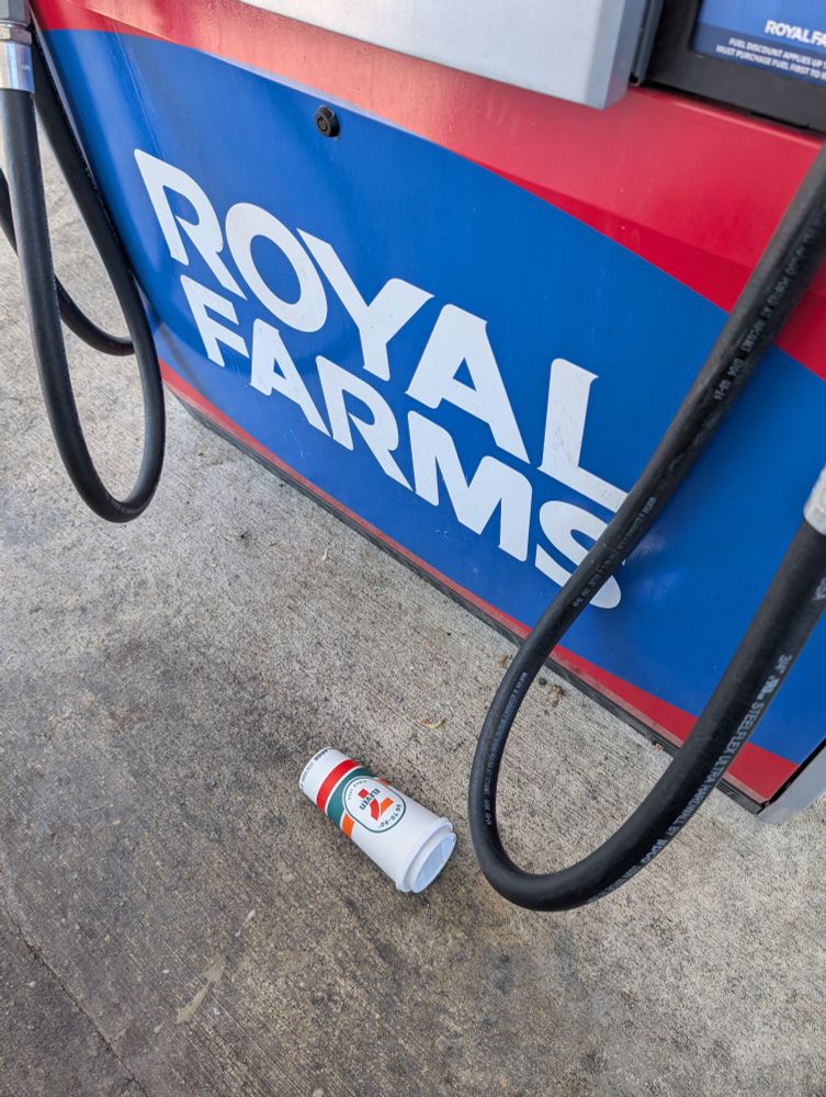 A picture of an empty 7-11 cup on the ground, beside a gas pump for Royal Farms