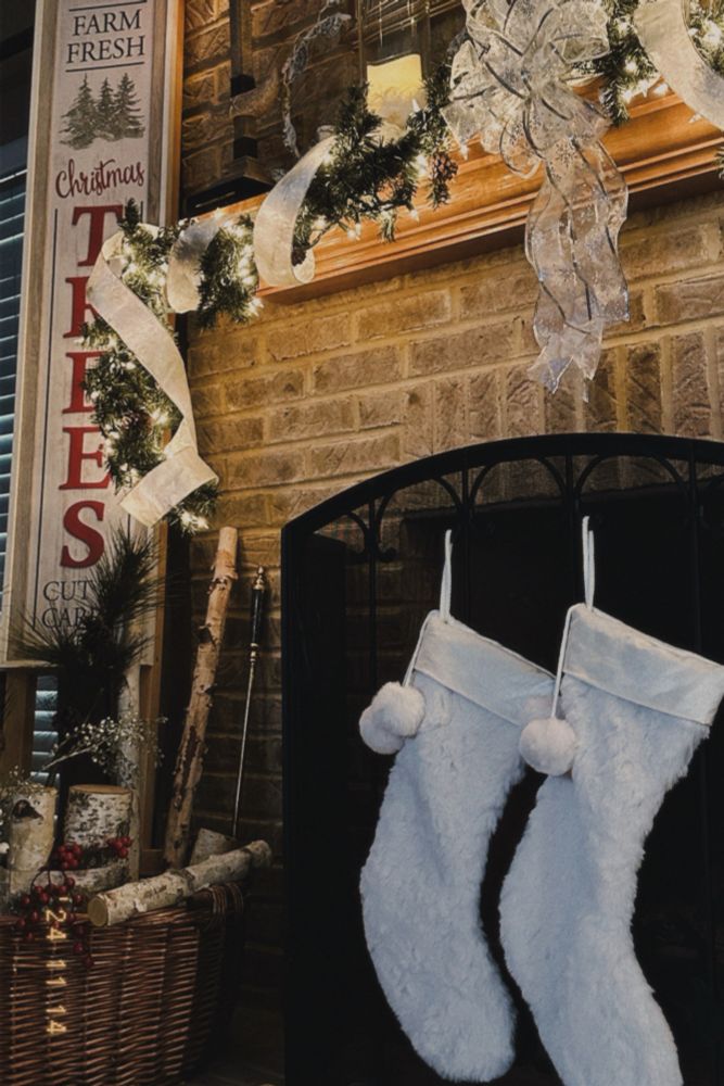 Two fluffy white stockings hanging by a brick fireplace below a mantle that is decorated with garland, string lights and ribbon.