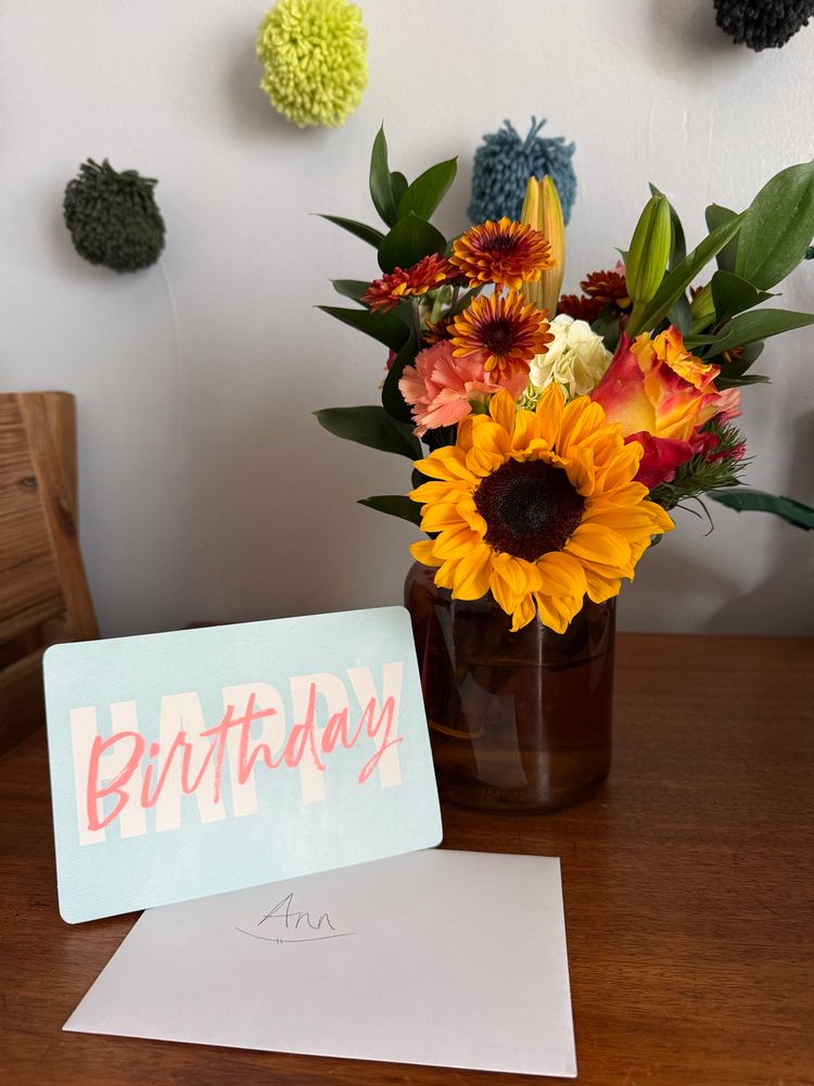 A bouquet of warm-toned flowers and greens in a brown vase sitting on a wood table next to a Happy Birthday card and an envelope with the name “Ann” written on it.