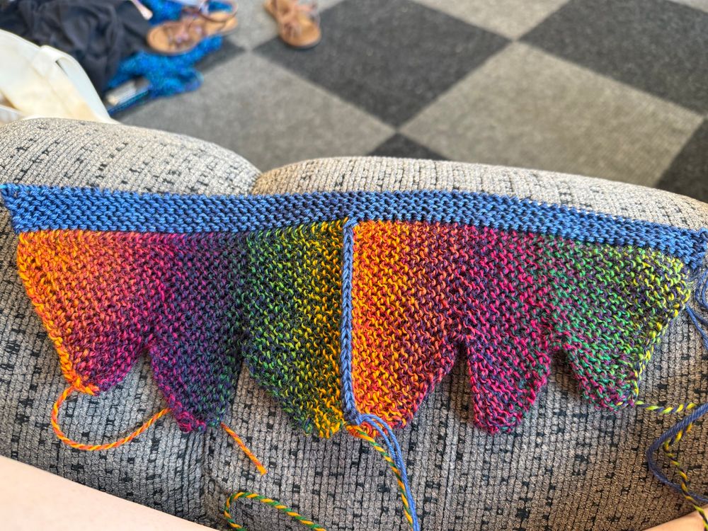 Flattened out tail section of a knitted emotional support chicken. A shift of rainbow hues oranges to purples to greens and back again. Edged with crayon blue. 