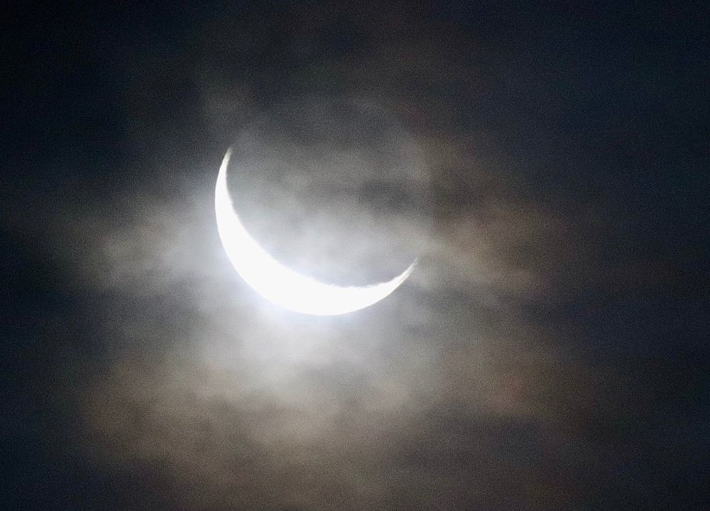 A crescent moon in early morning clouds in a photo that shows a vague outline of the entire moon.