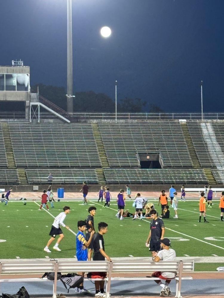 Picture of a full moon over a stadium with empty bleachers and a bunch of kids playing soccer on the field