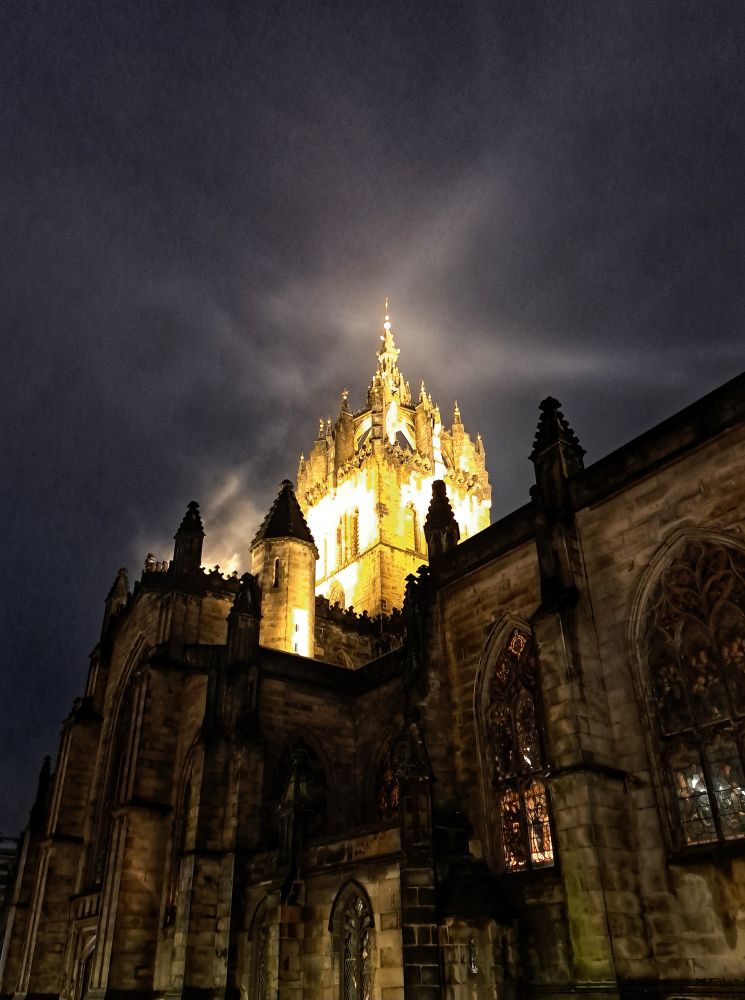 In the centre of a very dark image is the tower of a church, brightly lit with floodlights through a mist. The lower part of the church is not lit, barely visible. 