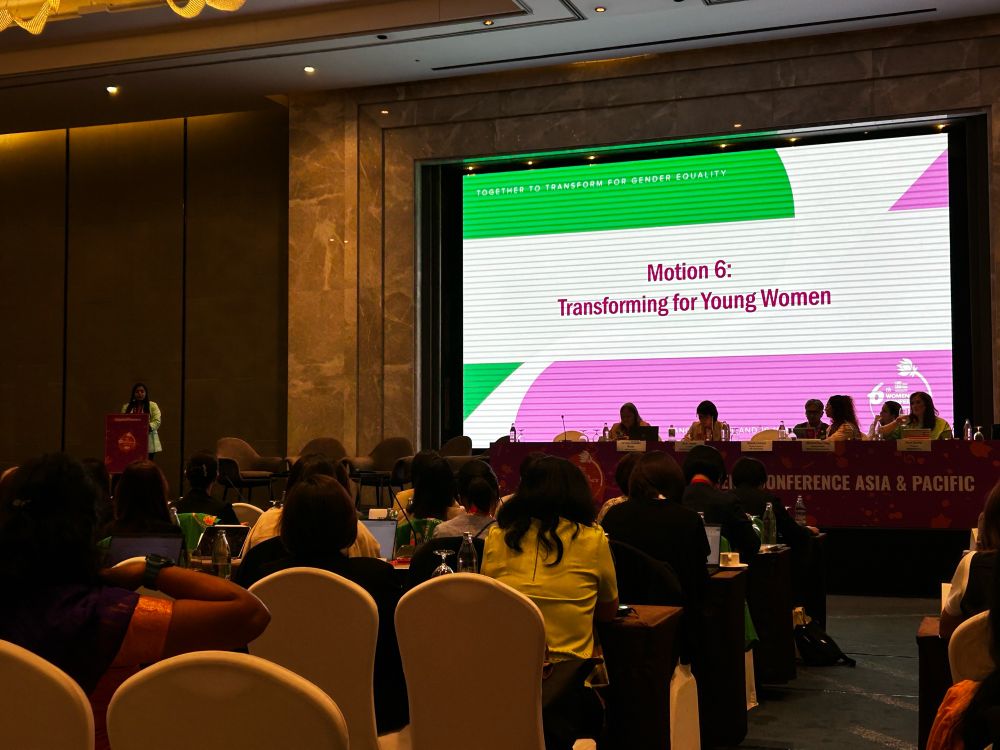 Photo shows speakers, panellists, and attendees at the UNI APRO Women’s Conference. The pink, green, and white projection on the big screen onstage says ‘Motion 6: Transforming for Young Women’. 