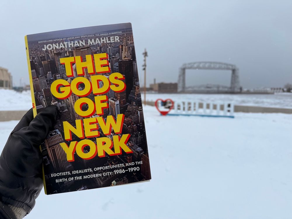 Black-gloved hand holds hardcover book, “The Gods of New York” by Jonathan Mahler, in front of snowy harbor with aerial lift bridge 