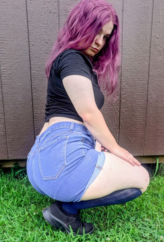 A photo of me posing, kneeling down with my body facing away from the camera, but I'm looking at the camera. my hair is a darker purple, my shirt is black, my shorts are denim, my shoes are black, and my knee highs are a dark blue. 