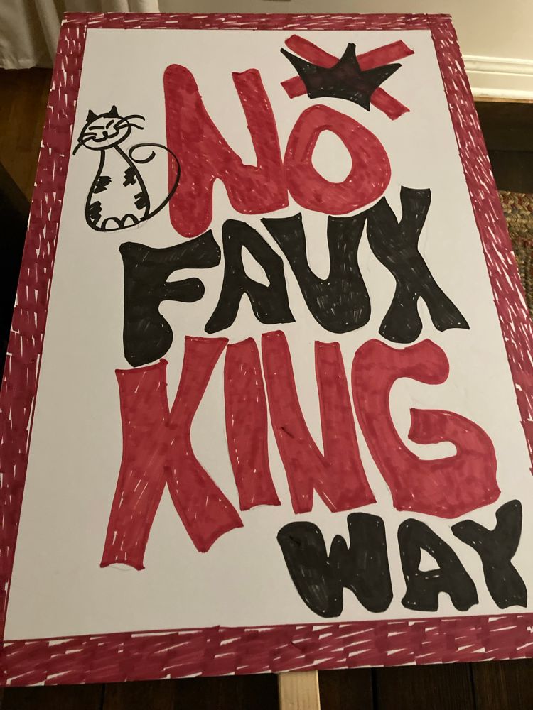 Front of protest sign reads: “No faux king way” with a crossed-out crown a drawing of a cat