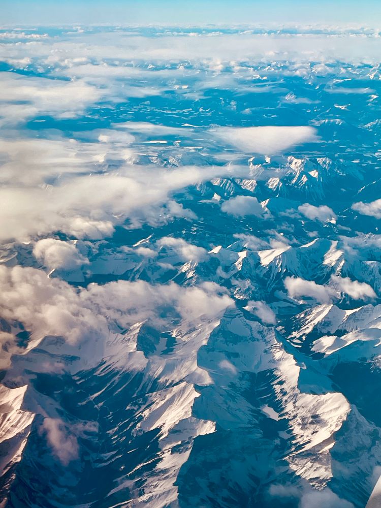 A photography taken from an airplane of the mountains lit by morning sun. They are white from snow on the left side and dark blue from shadow on the right. There are puffy white clouds floating over them. 