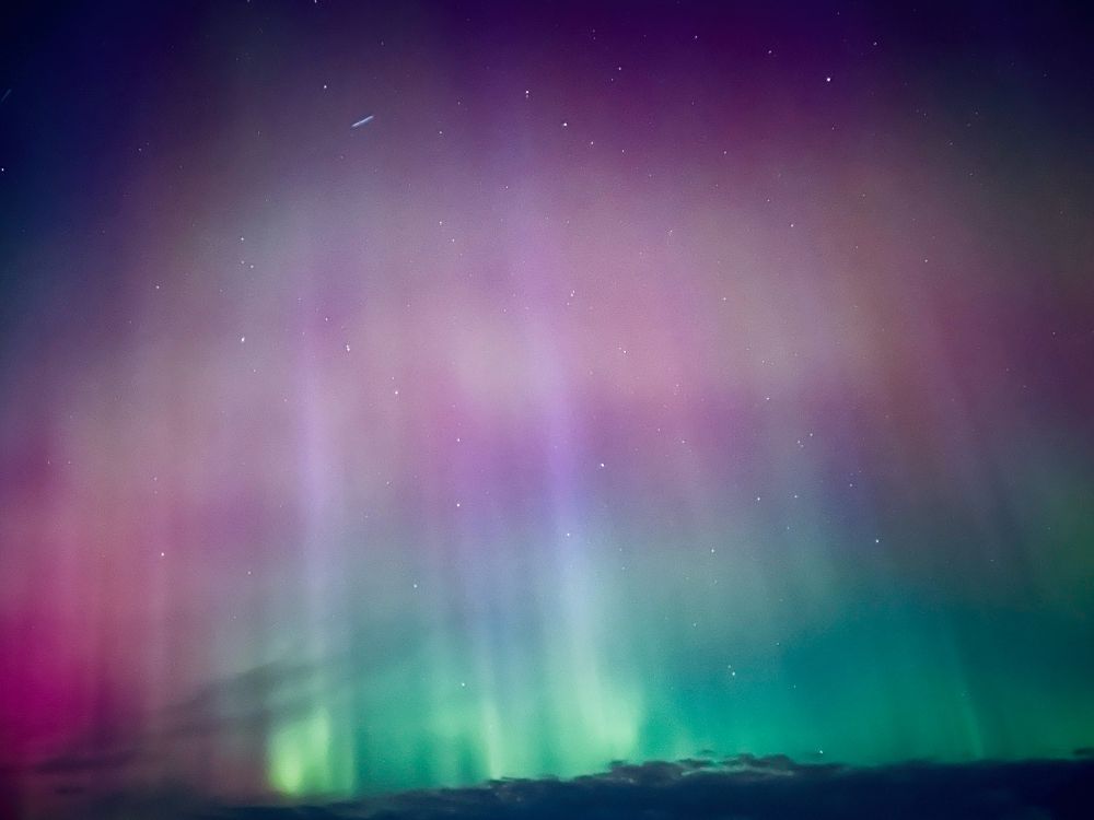 The northern lights above a home with blue, green, and pink in the sky. The lights are vertical with bright lines and stars. 