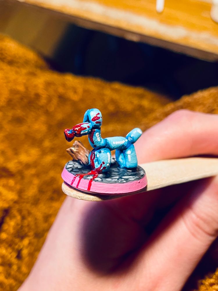 A miniature for DnD that is in the shape of a dog balloon animal form the mini is painted bright blue with white highlights and has red paint splattered on the face as if it were blood. The base is painted pink to represent the Trans flag. 