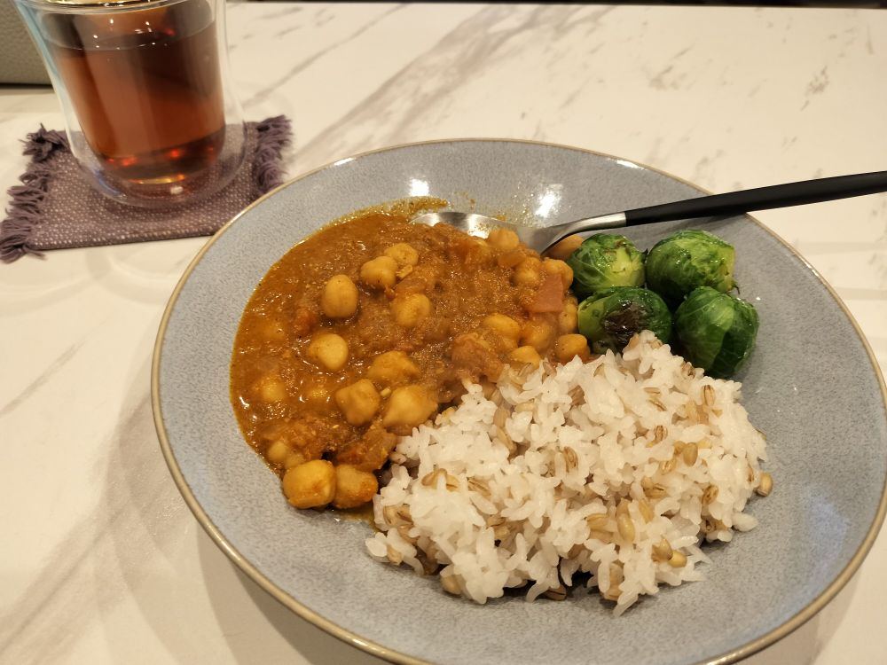 画面左上にお茶の入ったガラスのコップ、中央にチャナマサラと芽キャベツの入ったグレーのお皿。これらが白い食卓に置かれている。