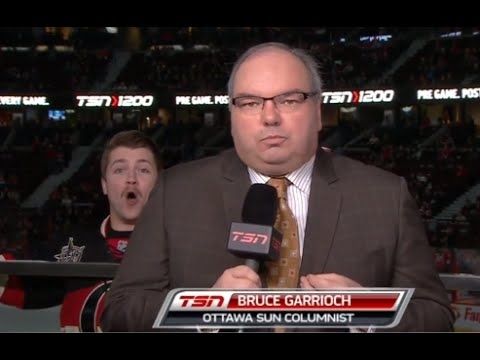 Ottawa Senators commentator Bruce Garrioch looking upset at broadcast camera at a game as fan behind is yelling