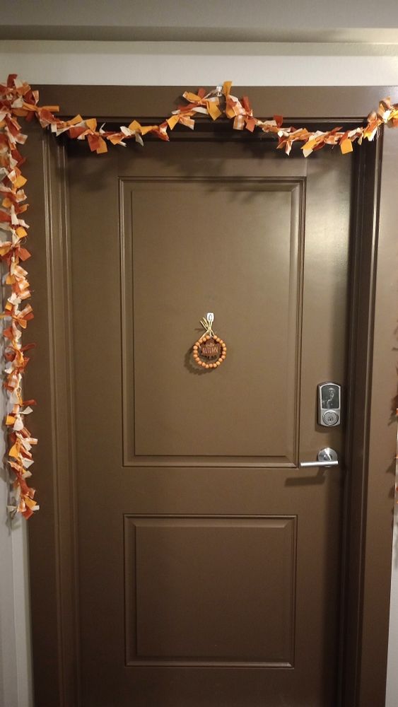 An apartment doorway decorated for fall. A garland with orange, white, and brown tassels drapes the door frame, and a wooden maple leaf hangs from the door.