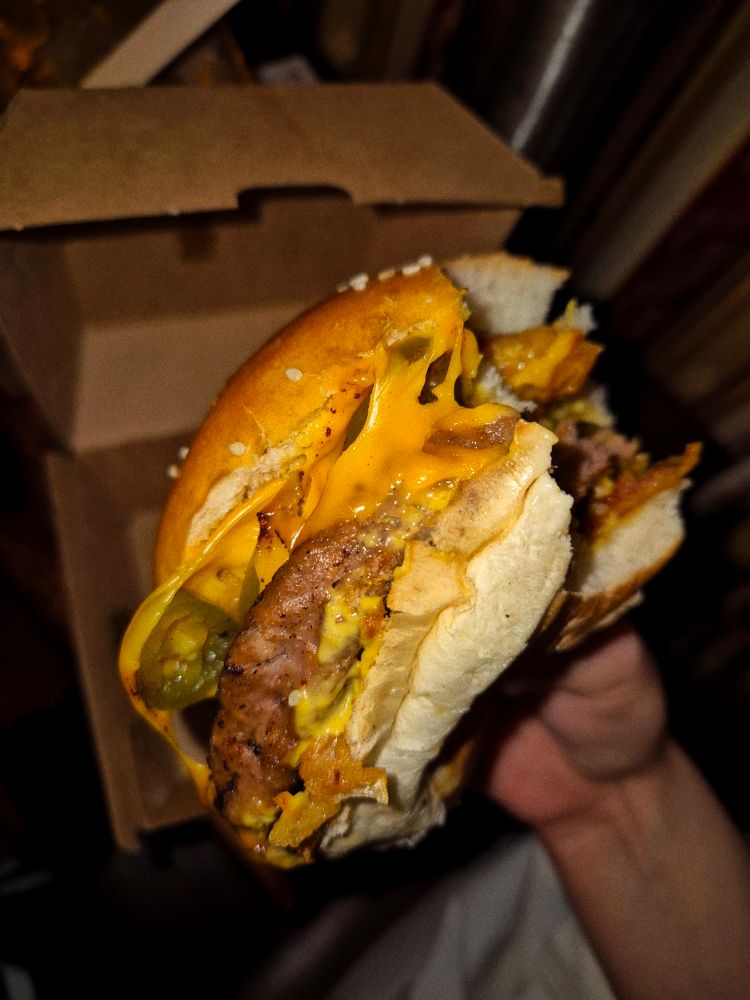 Man sieht meine Hand, die einen großen Burger hält. Auf dem Burger befinden sich ein Patty, veganer Käse, Jalapenos, Sauce und Zwiebelringe.