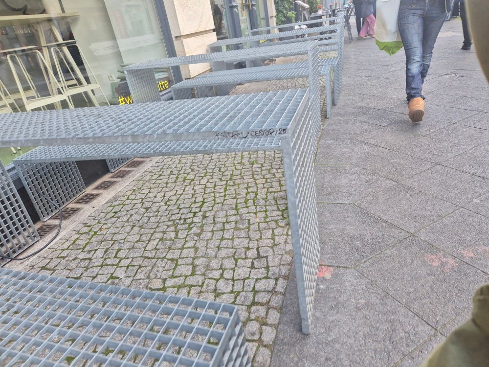 Foto im Querformat. Zu sehen ist der Außenbereich eines Restaurants neben einer Fußgänger*innenzone. Vor dem Restaurant stehem Bänke und Tische aus Metallgitter ohne Kissen.
