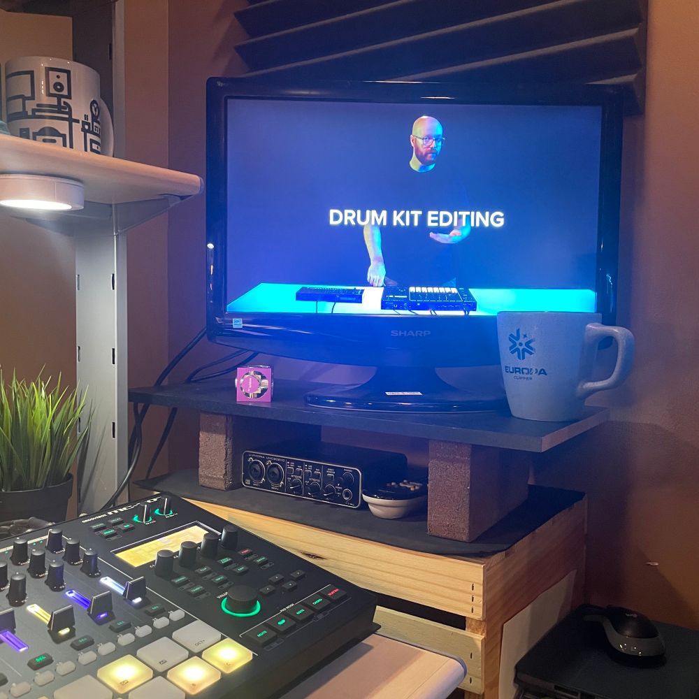 Computer monitor on a table with a drinking mug and groovebox controller on a desk, with a faux plant.