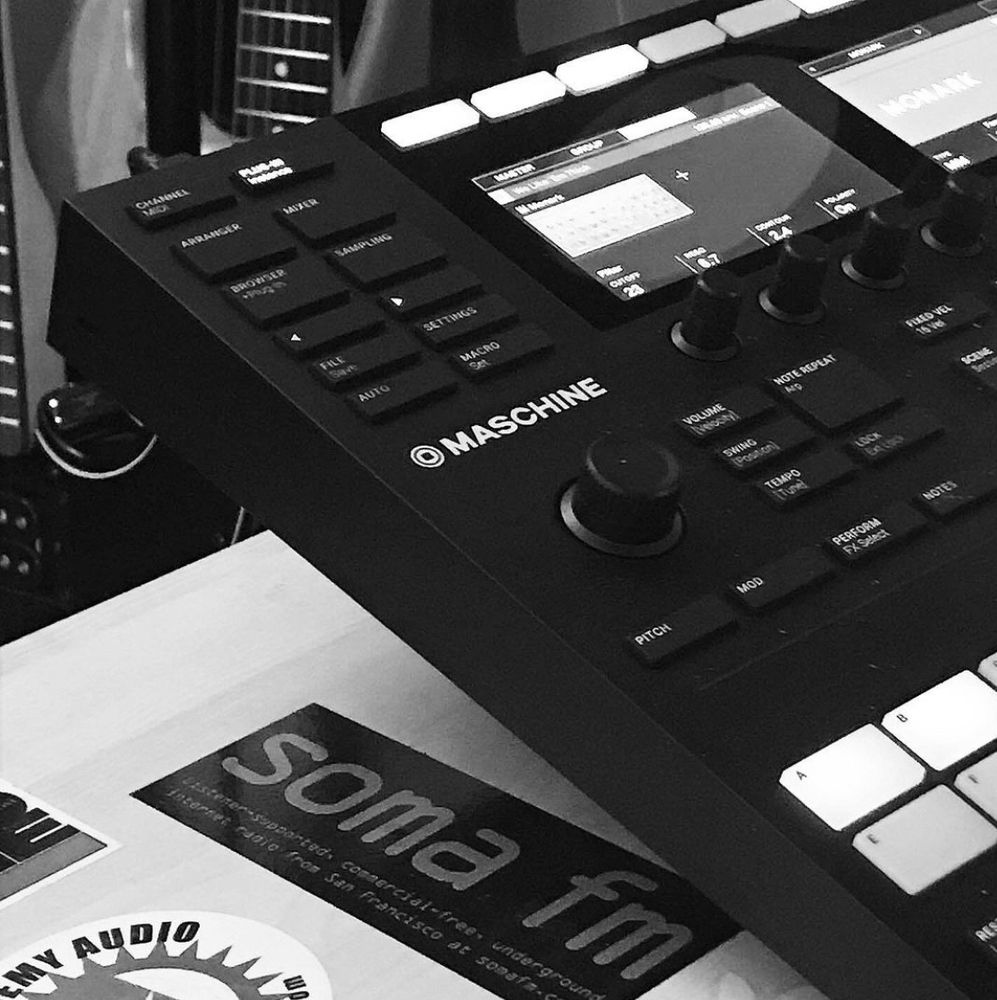 B&W photo : Native Instruments Maschine on a table, electric guitars in the background.