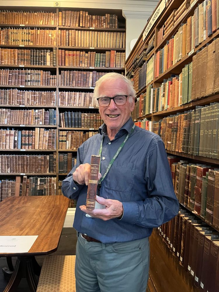 Library volunteer standing in a historic library holding a book called ‘The common sense of the exact sciences’.